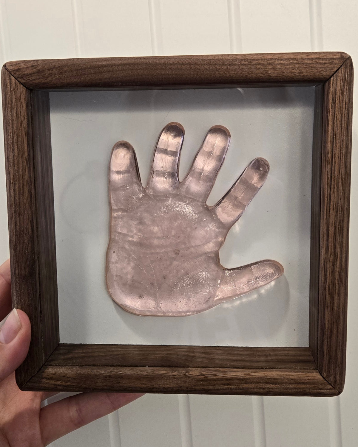 A hand holds a square dark wood display frame containing a translucent pink hand-shaped glass piece mounted on a light background.