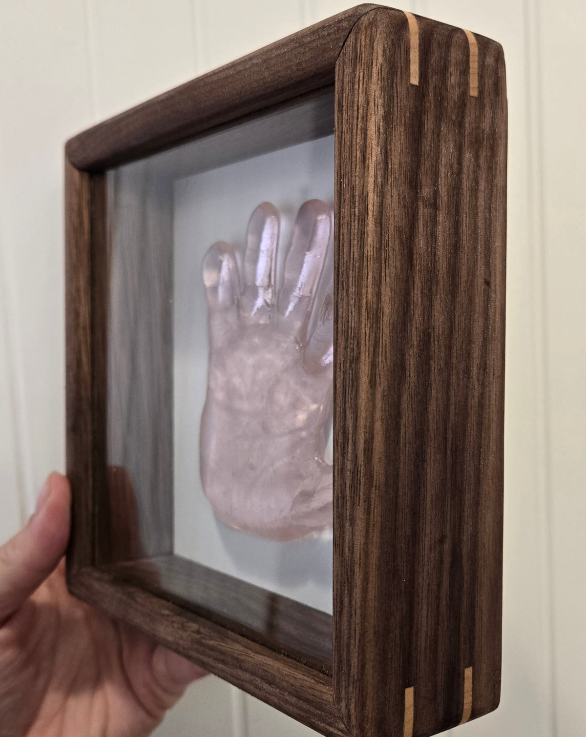 A hand holding a dark wood shadow box display featuring a light pink glass handprint keepsake mounted inside a glass-front frame.