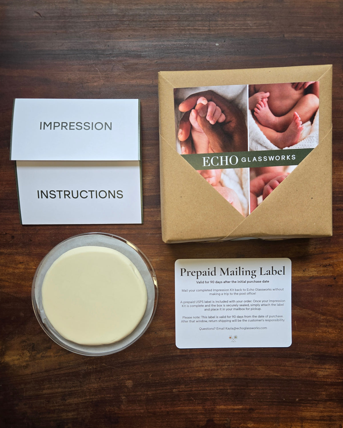 A baby print keepsake gift set from Echo Glassworks arranged on a dark wood background, including an instructions card, a round glass dish with pale yellow impression compound, and a prepaid mailing label next to a kraft gift box with baby footprints photos.