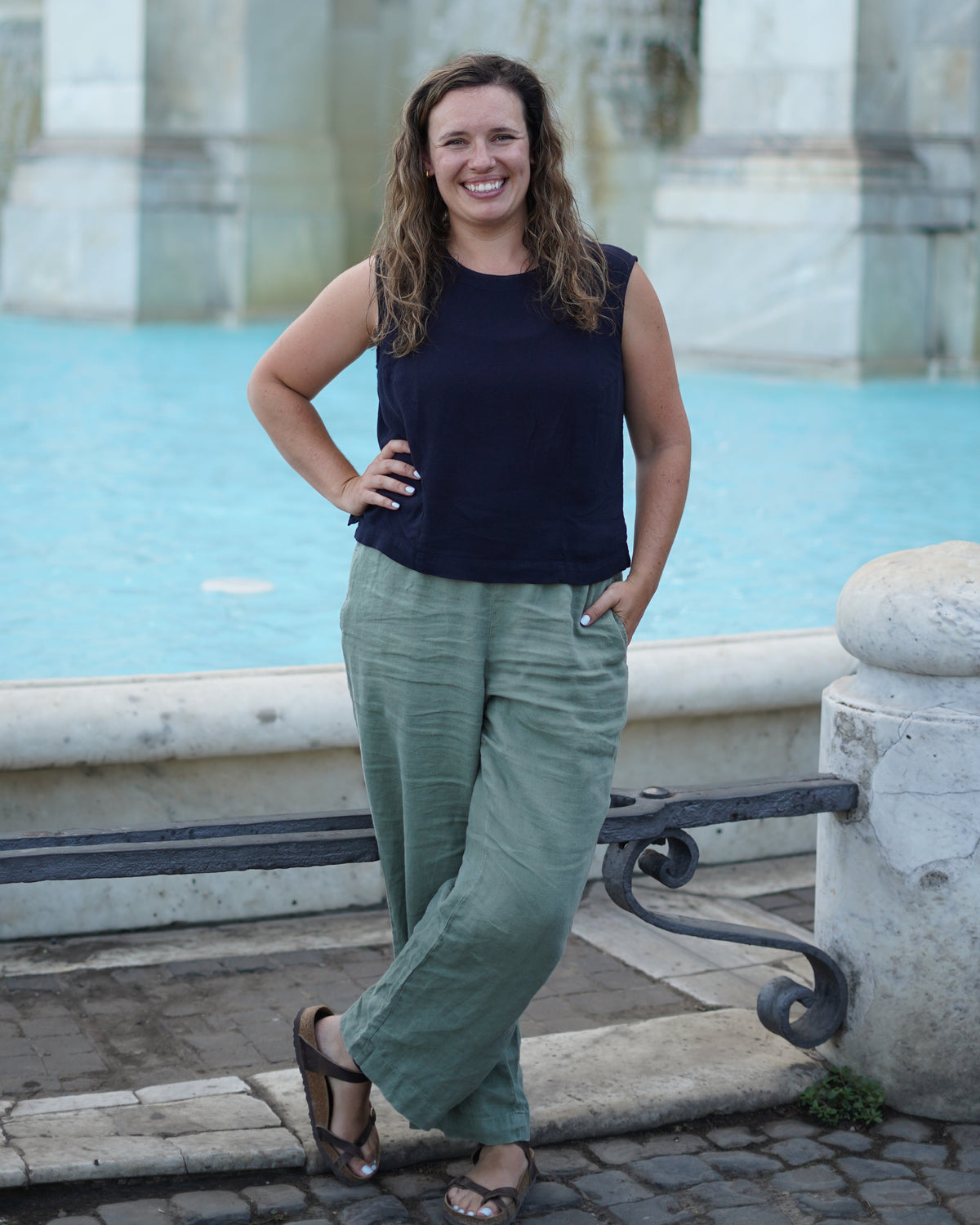 Brunette woman in back tank top and sage green pants stands with a pool of blue water and marble pillars in the background