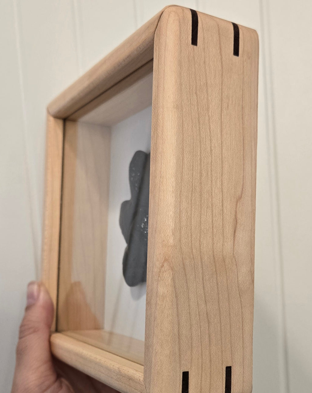 A close-up of a hand holding a square, light natural wood shadow box frame with a clear front panel and a black glass keepsake set against a white paneled wall.