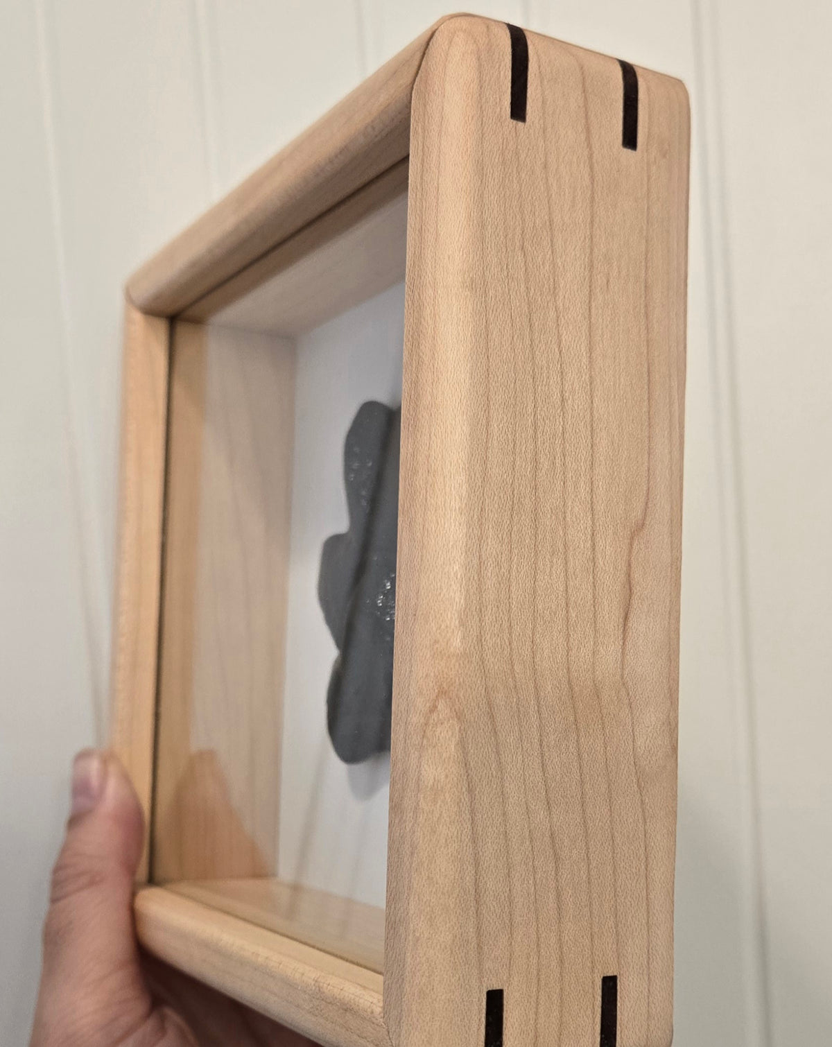 A close-up of a hand holding a square, light natural wood shadow box frame with a clear front panel and a black glass keepsake set against a white paneled wall.