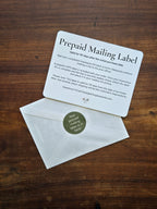 A prepaid mailing label card with printed instructions lies on top of a white envelope on a dark wood surface. The envelope has a round green sticker that reads, 'Your prepaid mailing label is here!'.