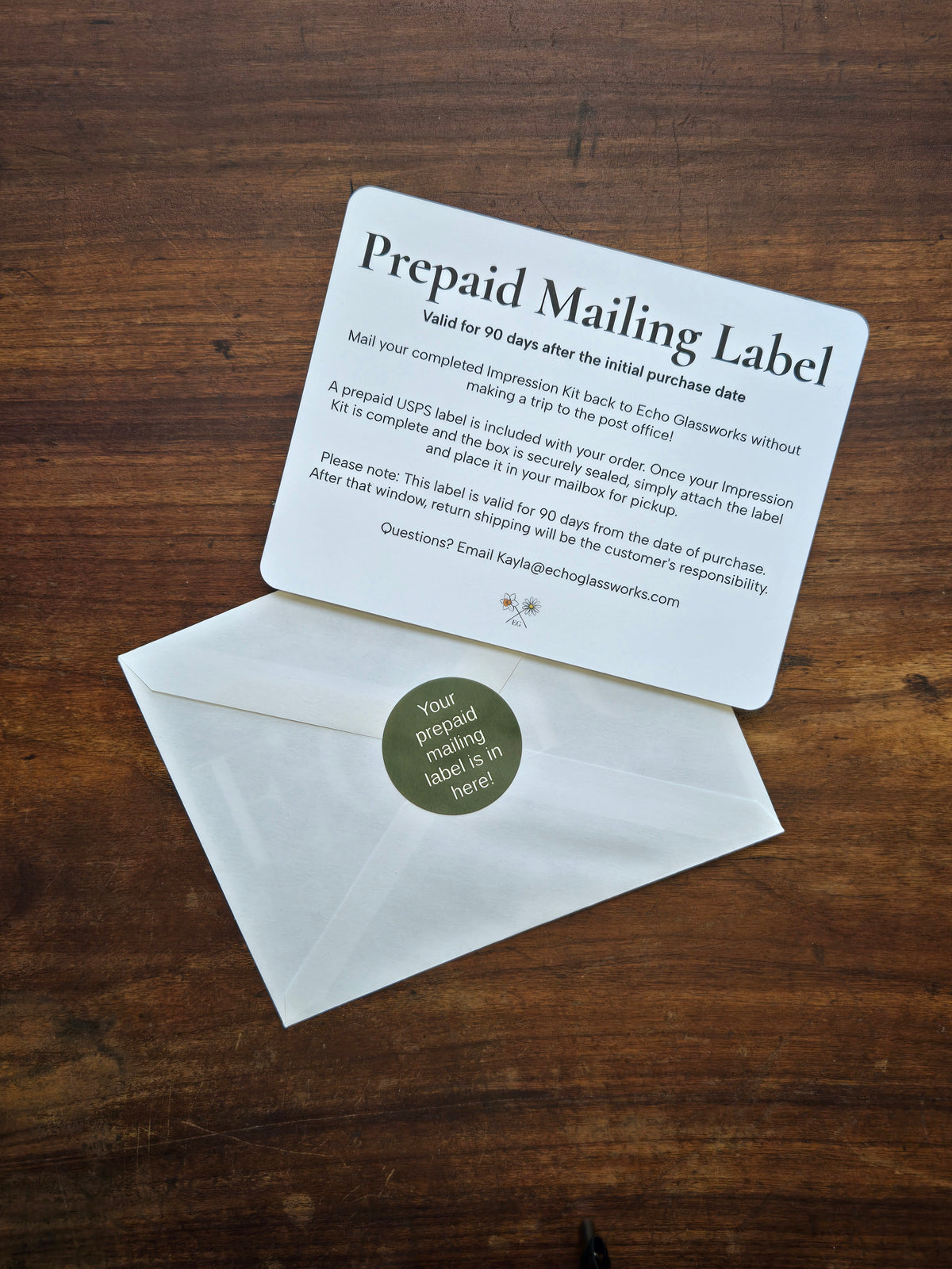 A prepaid mailing label card with printed instructions lies on top of a white envelope on a dark wood surface. The envelope has a round green sticker that reads, 'Your prepaid mailing label is here!'.