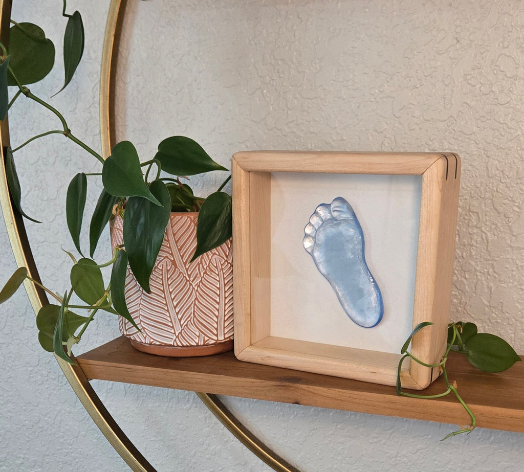 A wooden shadow box frame displaying a blue glass footprint, styled on a wooden shelf next to a potted plant with a gold circular frame against a textured beige wall.