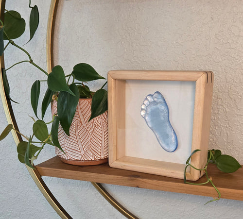 A wooden shadow box frame displaying a blue glass footprint, styled on a wooden shelf next to a potted plant with a gold circular frame against a textured beige wall.