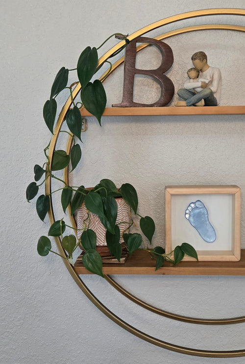 Half a circular wall shelf featuring a blue glass footprint in a shadowbox surrounded by various shelf items: a green plant, a father-child sculpture, and a wooden letter "B"