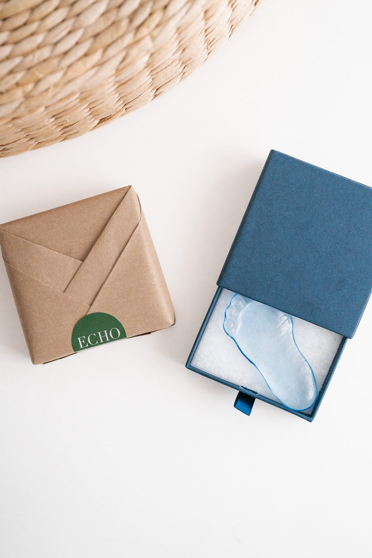A light blue glass footprint keepsake displayed in an open dark blue presentation box next to a brown kraft paper envelope-style package with a green circular 'ECHO' logo, on a white background with a woven mat in the corner.