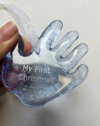 An example of an engraved handprint from Echo Glassworks: A hand holding a translucent blue glass Christmas ornament shaped like a hand, with the text 'My First Christmas' engraved on it and a white ribbon loop.