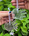 Two adult hands hold two glass baby handprint replicas hanging from ribbons on a green leafy background