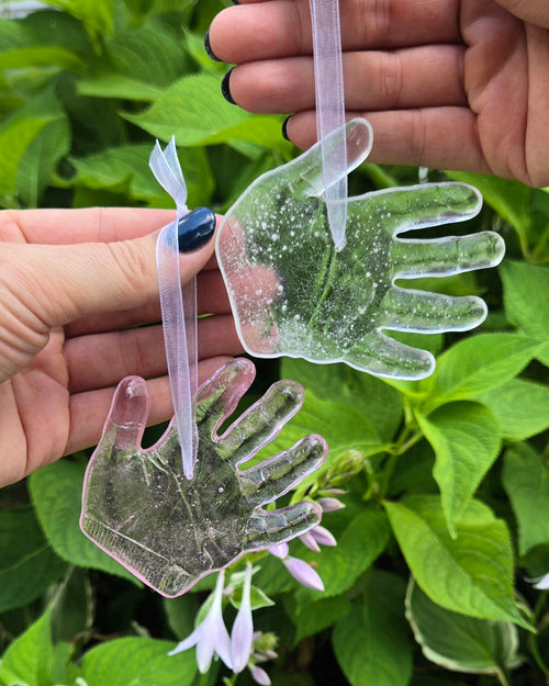 Two adult hands hold two glass baby handprint replicas hanging from ribbons on a green leafy background