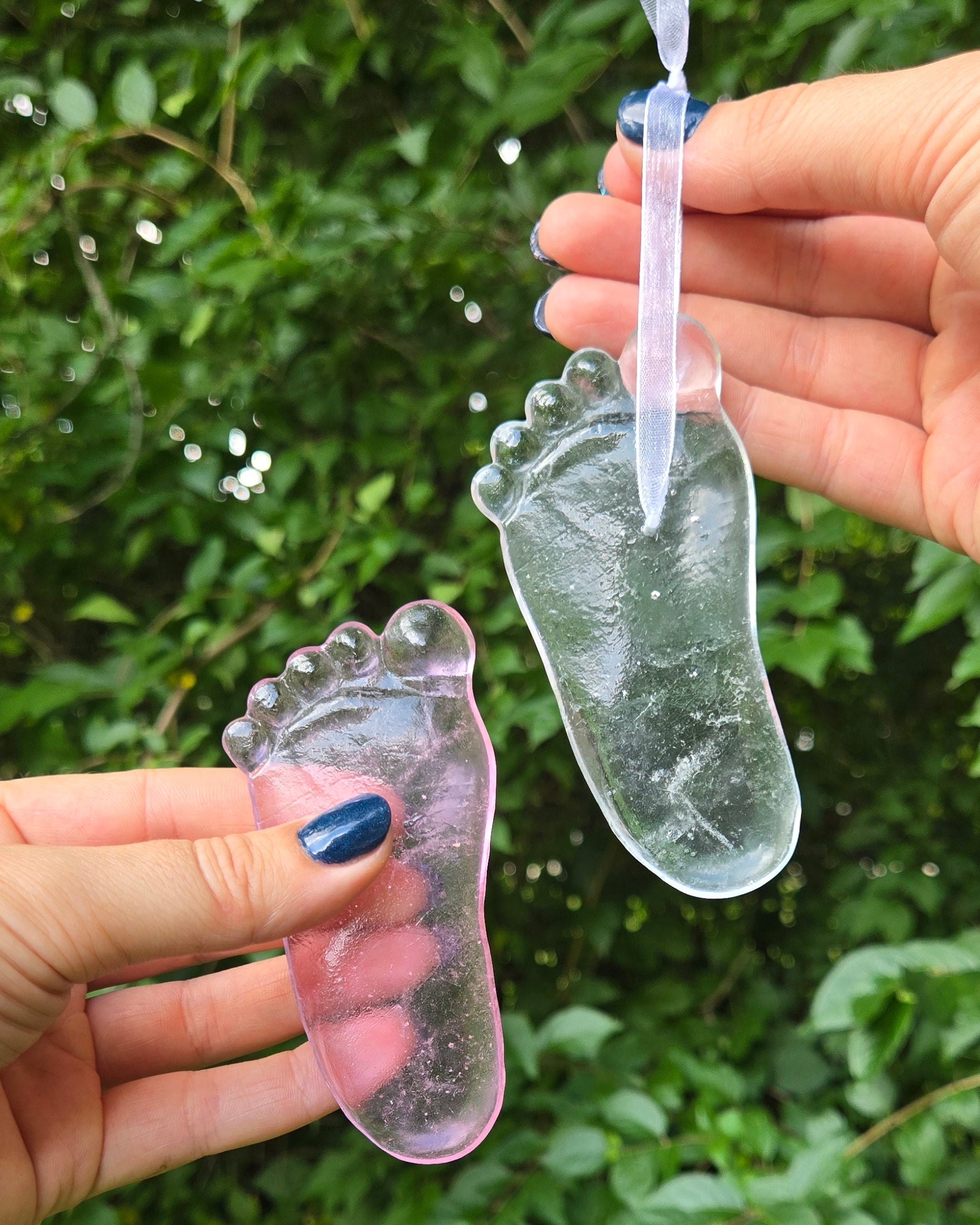 Two adult hands hold two glass baby footprint replicas on a leafy green background