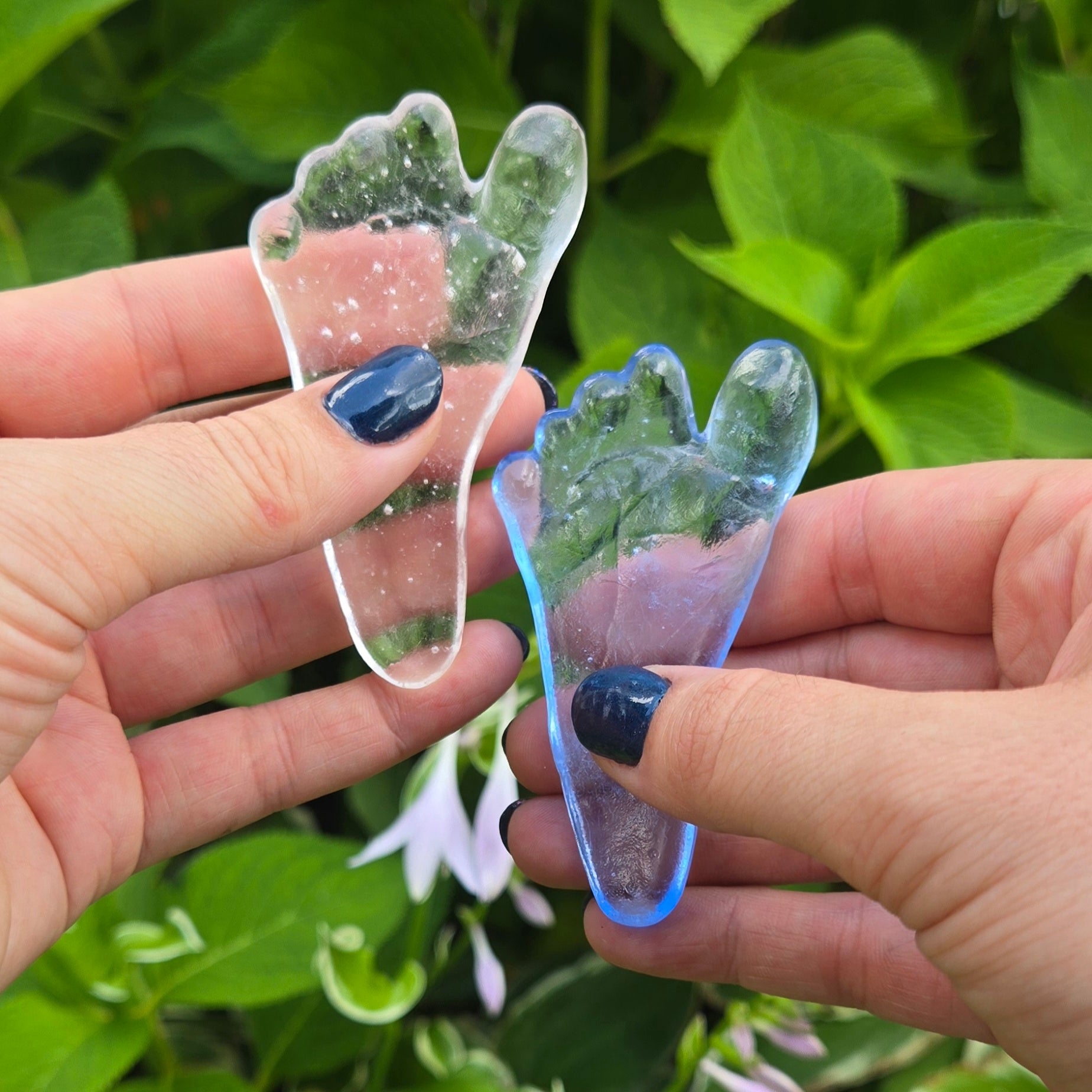 Two adult hands hold two glass baby footprint replicas on a leafy green background